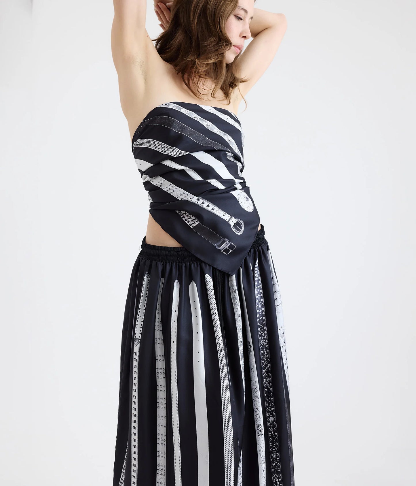 Woman wearing a black and white silk scarf with images of belts as a top and a black and white silk skirt with the same belt print standing in front of a white background