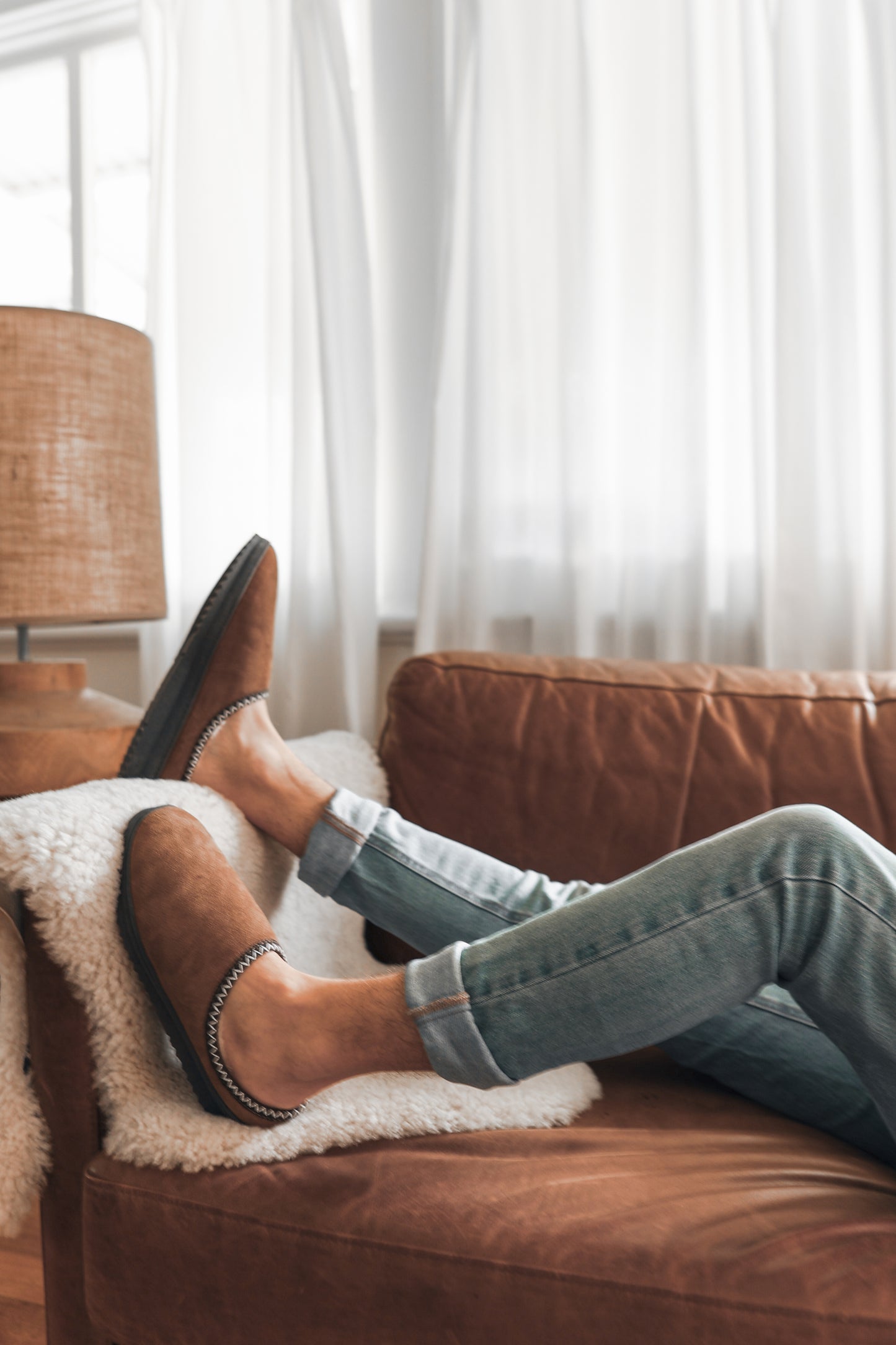 Person wearing brown short uggs and jeans lounging on a brown leather couch with white curtains in the background.