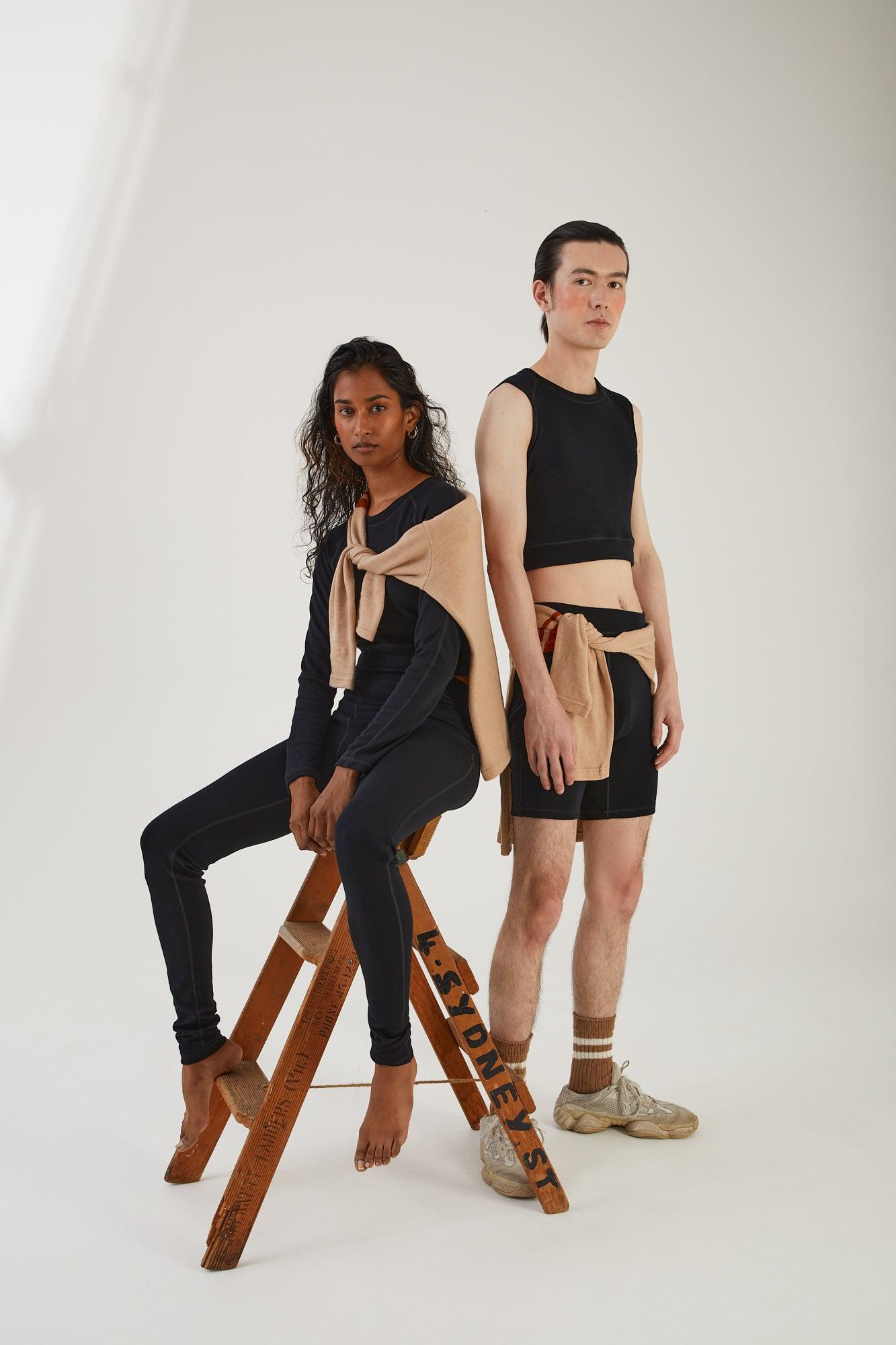 A person sitting on a wooden step ladder wearing merino wool leggings and a merino wool cropped long sleeve top with a merino wool jumper draped around their shoulders next to a person standing wearing merino wool socks, merino wool biker shorts and merino wool cropped tank with a merino wool jumper tied around their waist