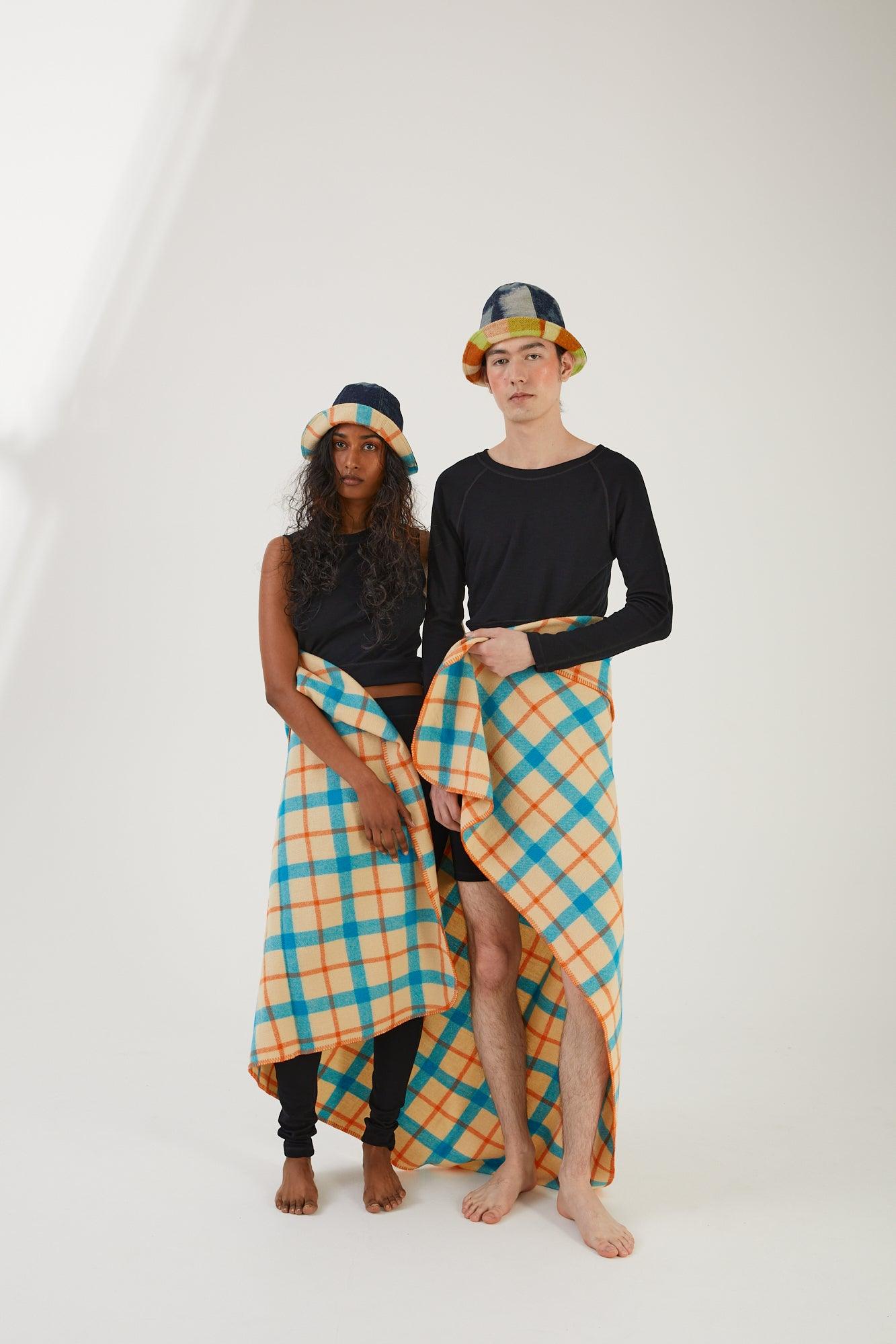 Two people standing side by side with a wool throw wrapped around their waists and upcycled hats. One person is wearing merino wool leggings and a merino wool tank, the other is wearing merino wool biker shorts and a merino wool cropped tank
