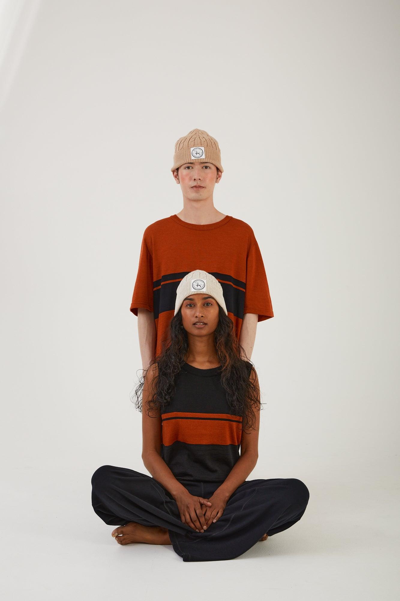 A person wearing a merino wool top and a merino wool beanie with someone sitting directly in front of them with their legs crossed wearing a merino wool beanie, merino wool vest and merino wool track pants