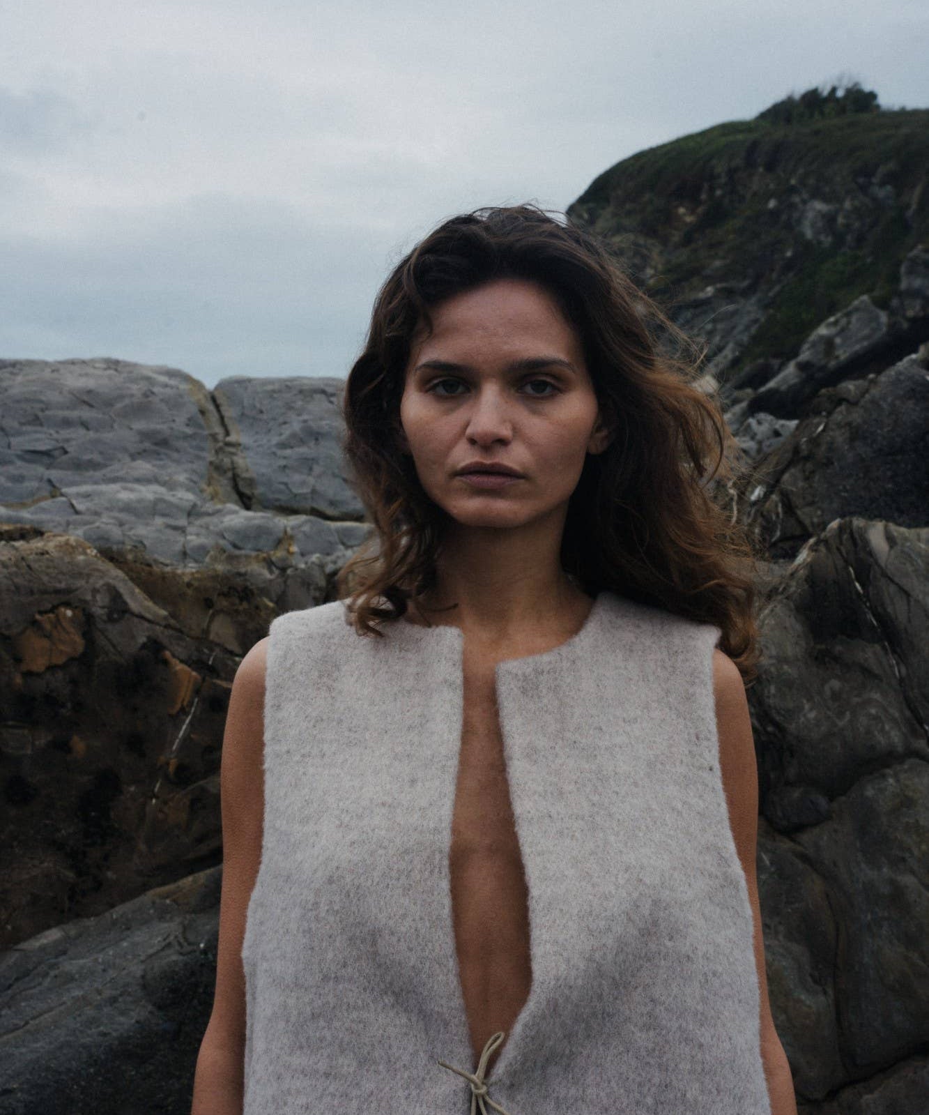 Woman wearing a sleeveless beige vest standing against a rocky background