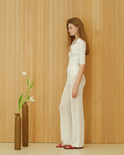 Person in a white outfit standing next to a vase with flowers against a wooden panelled wall.