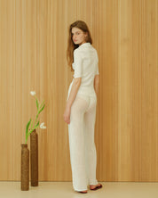 Person in a white outfit standing against a wooden panelled wall with two vases and flowers on the floor.