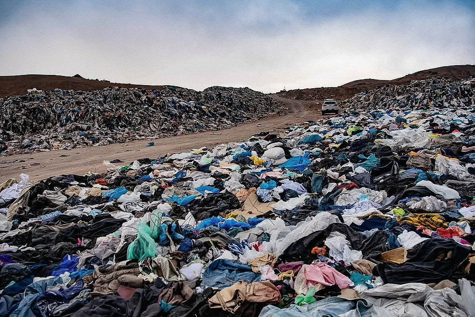 Large pile of discarded clothing at a landfill site with mountains in the background.