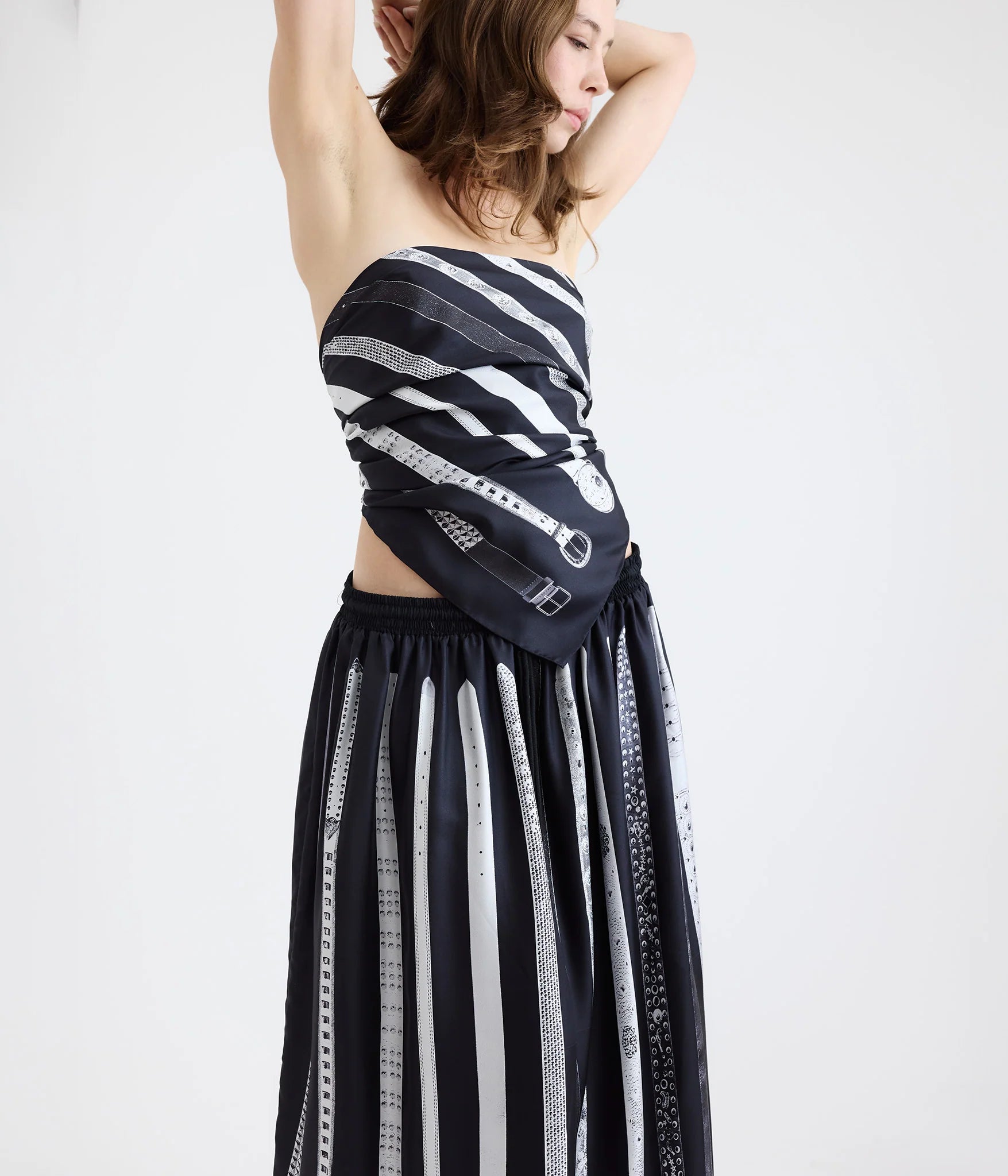 Woman wearing a black and white silk scarf with images of belts as a top and a black and white silk skirt with the same belt print standing in front of a white background