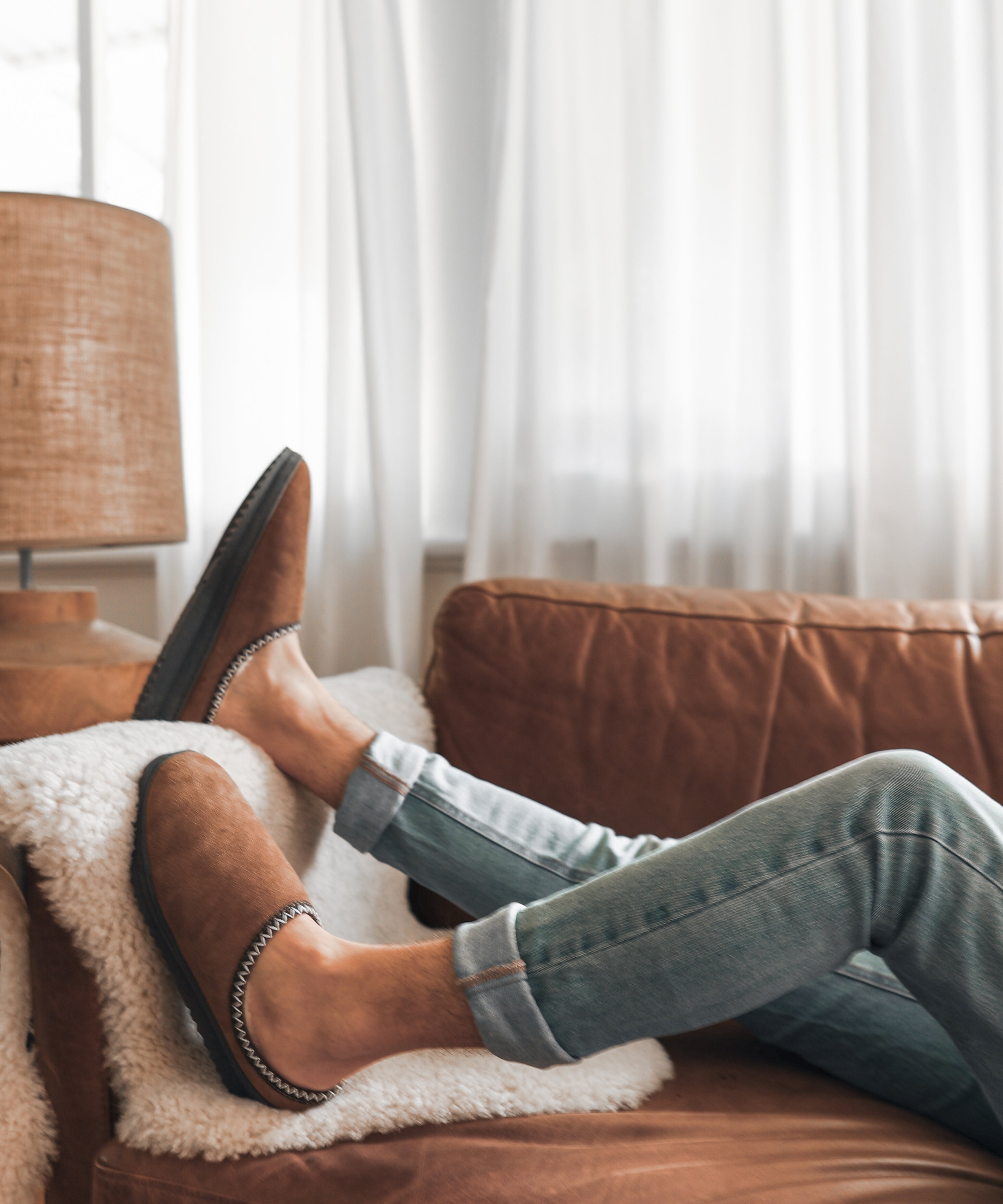 Person wearing brown short uggs and jeans lounging on a brown leather couch with white curtains in the background.