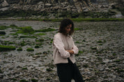 Woman standing on a rocky beach with a mountainous background in a beige vest