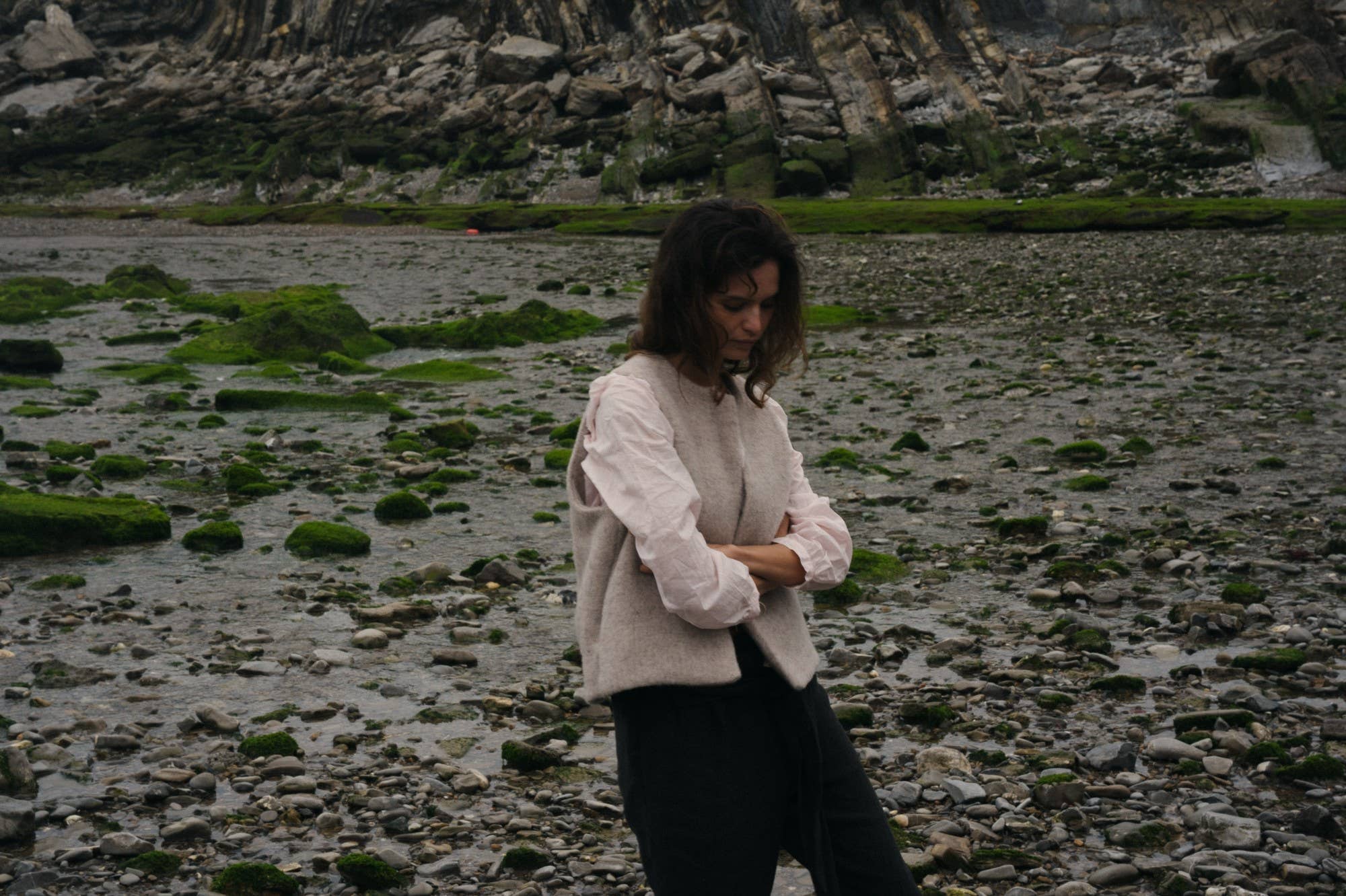 Woman standing on a rocky beach with a mountainous background in a beige vest