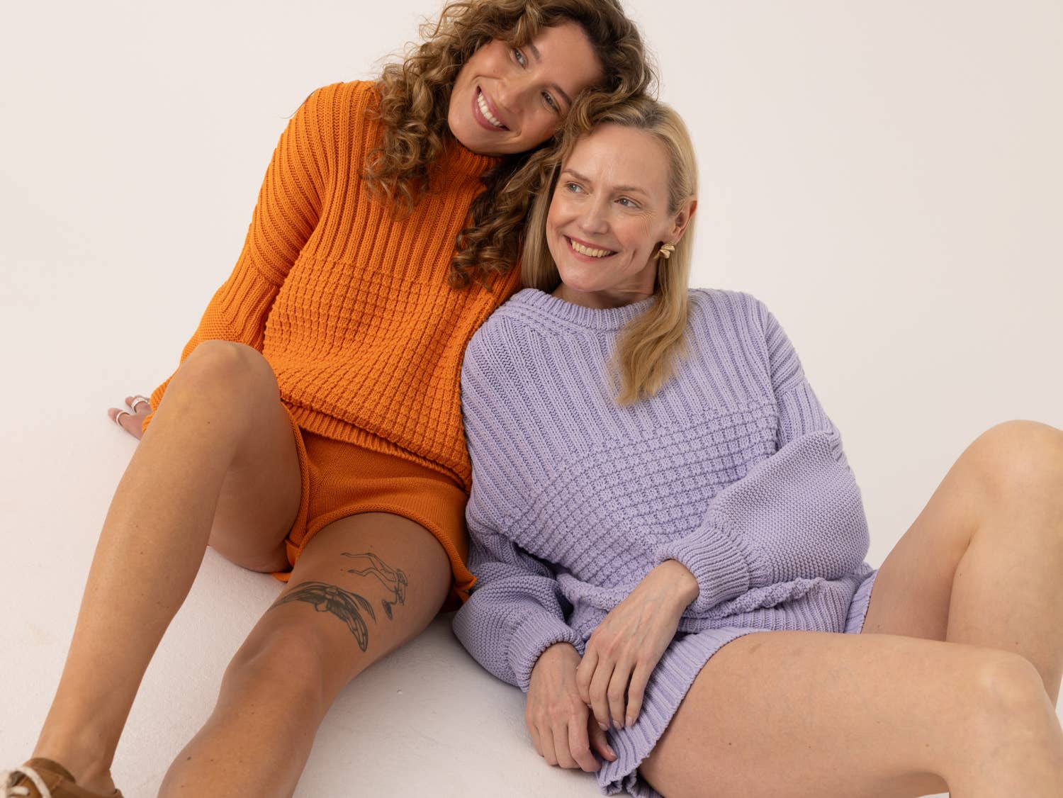 Two women sitting together on a white surface, wearing orange and purple knitted jumpers and shorts.