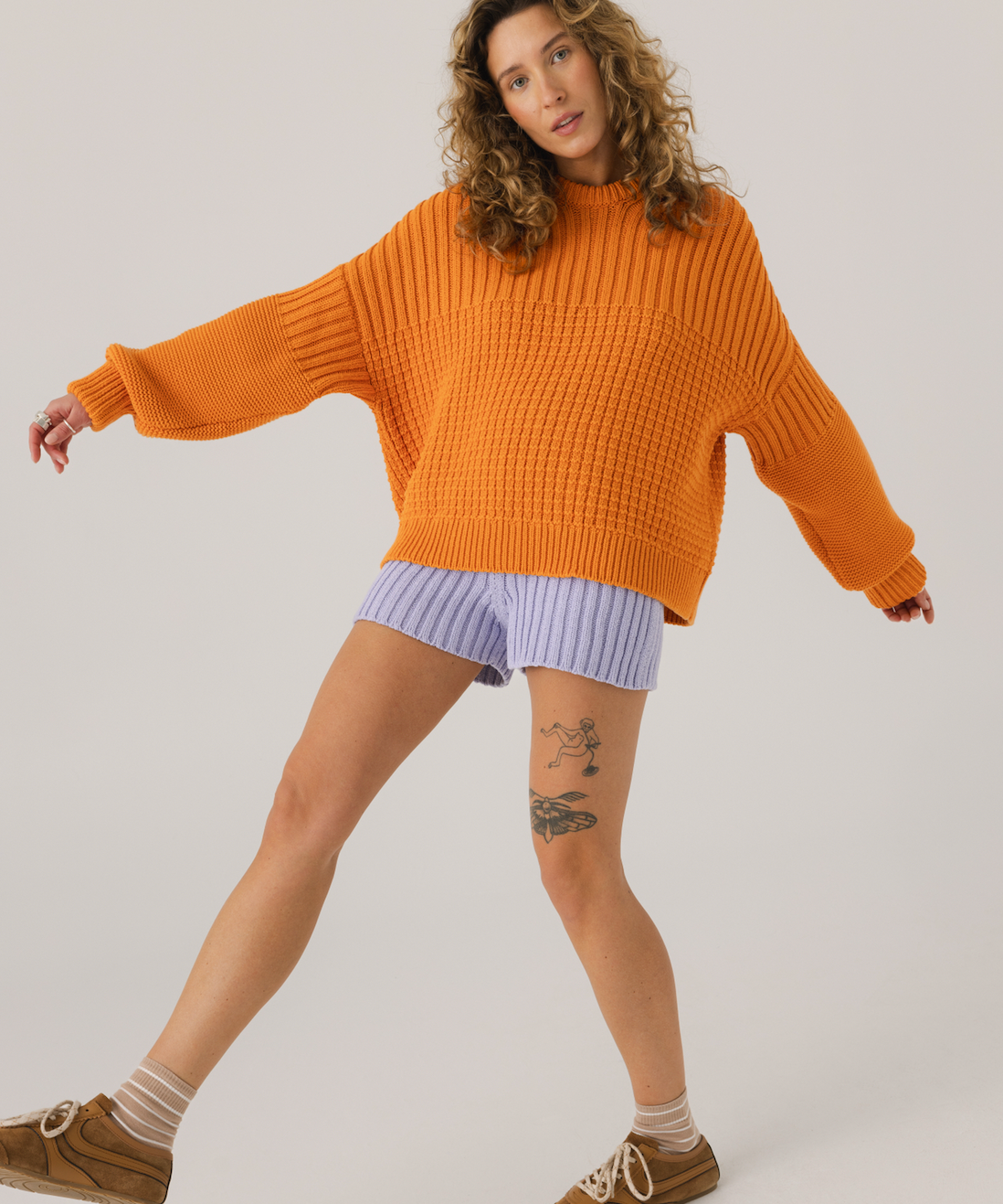 Woman wearing an orange sweater and light purple shorts on a plain background