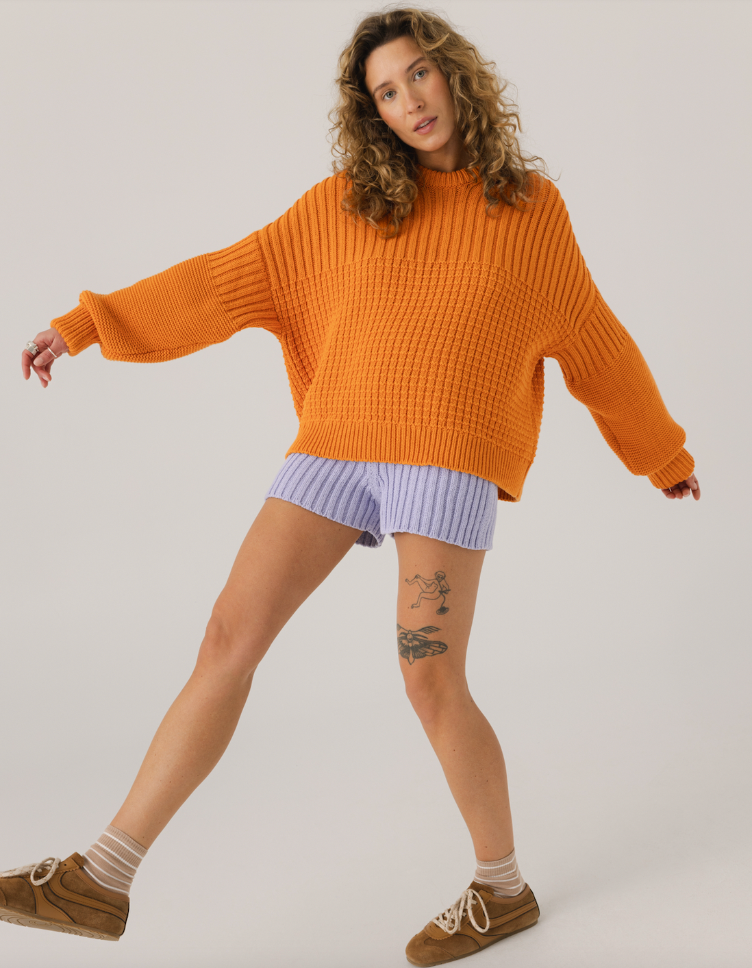 Woman wearing an orange sweater and light purple shorts on a plain background