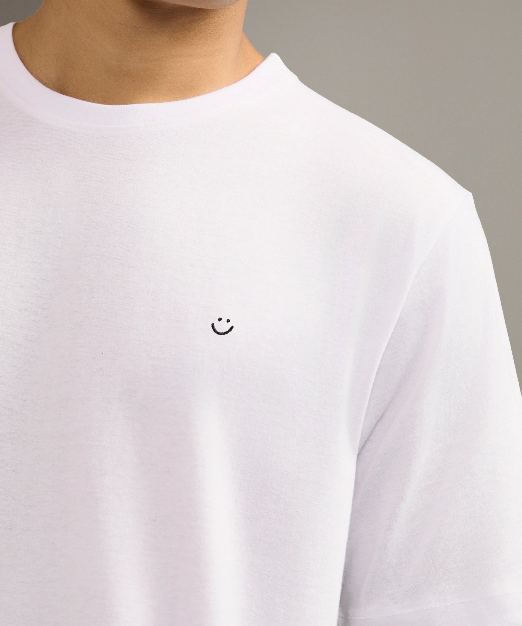 White t-shirt with a small smiley face logo worn by a person on a neutral background