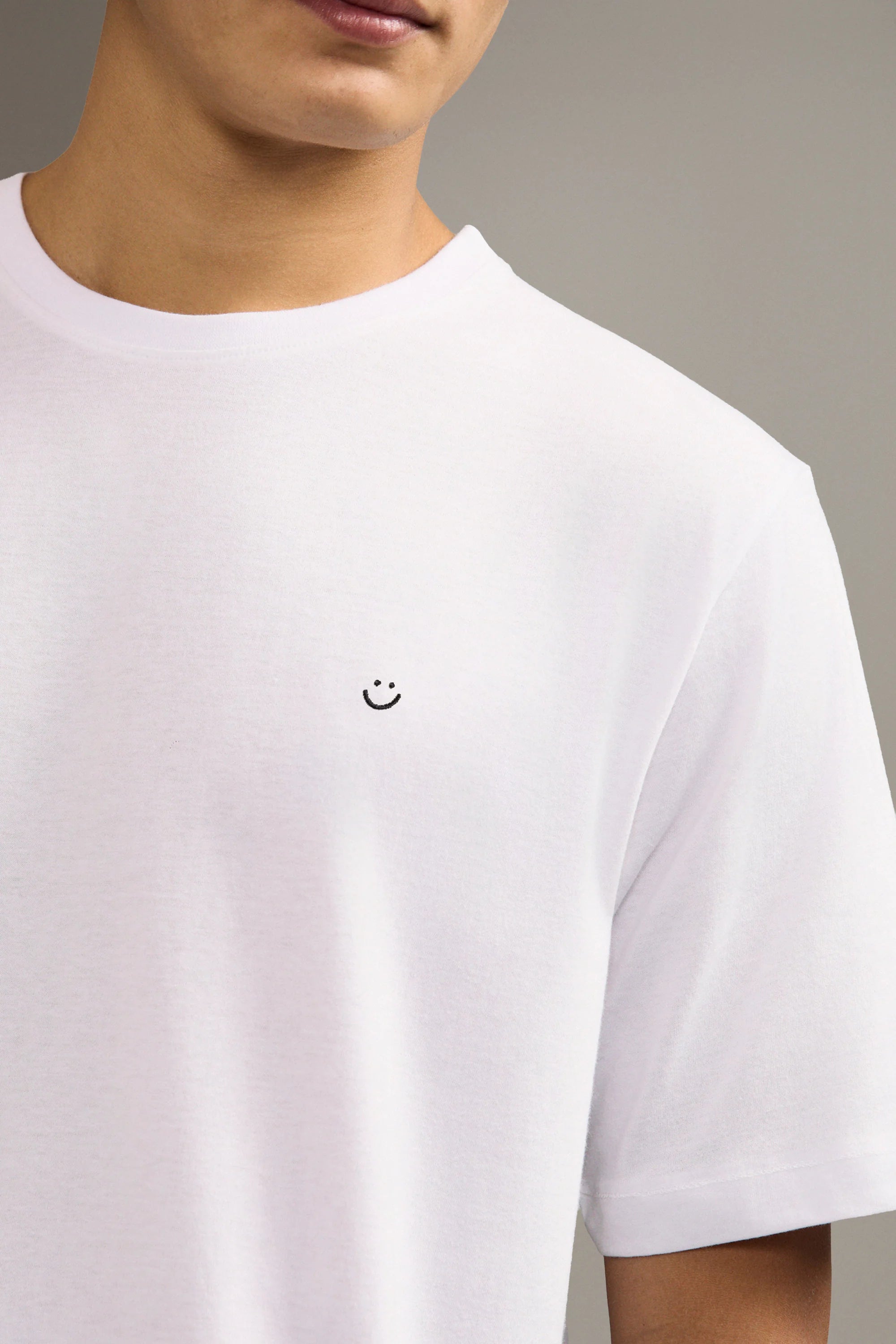 White t-shirt with a small smiley face logo worn by a person on a neutral background