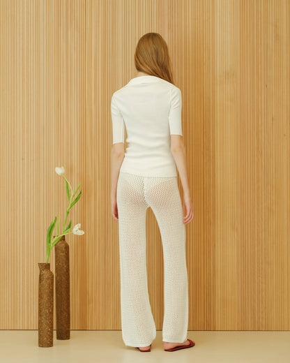 Person wearing a white knit outfit standing against a wooden panelled wall with two vases and a plant.