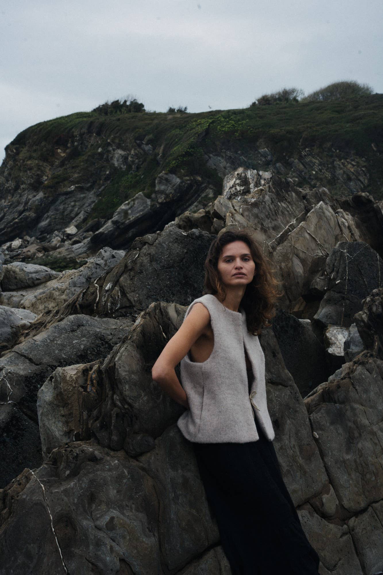 Woman standing on rocky terrain with a mountainous background in a beige vest