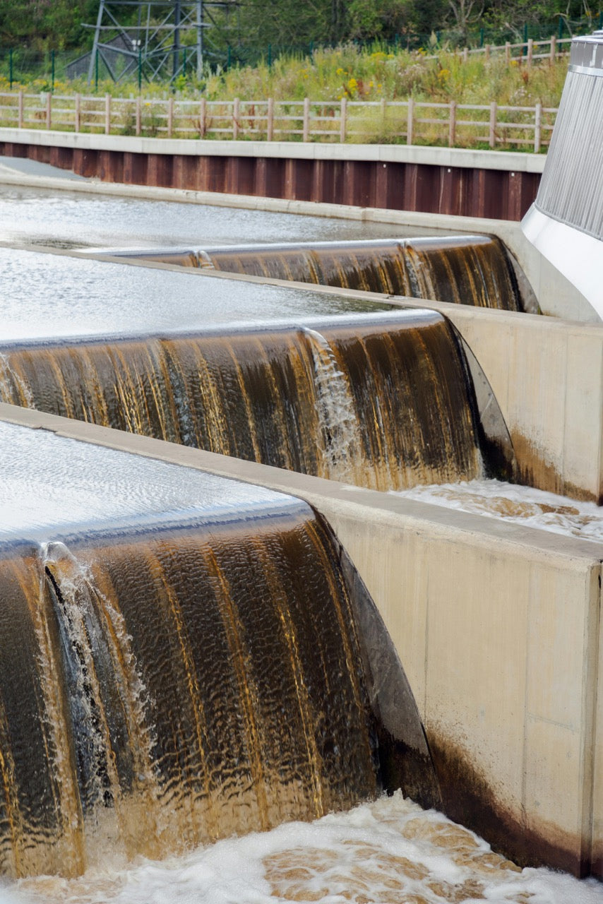 Water flowing over a series of concrete barriers with a natural landscape in the background.