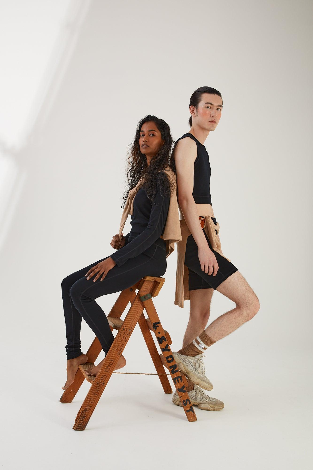 A person sitting on a wooden step ladder wearing merino wool leggings and a merino wool cropped long sleeve top with a merino wool jumper draped around their shoulders next to a person standing wearing merino wool socks, merino wool biker shorts and merino wool cropped tank with a merino wool jumper tied around their waist