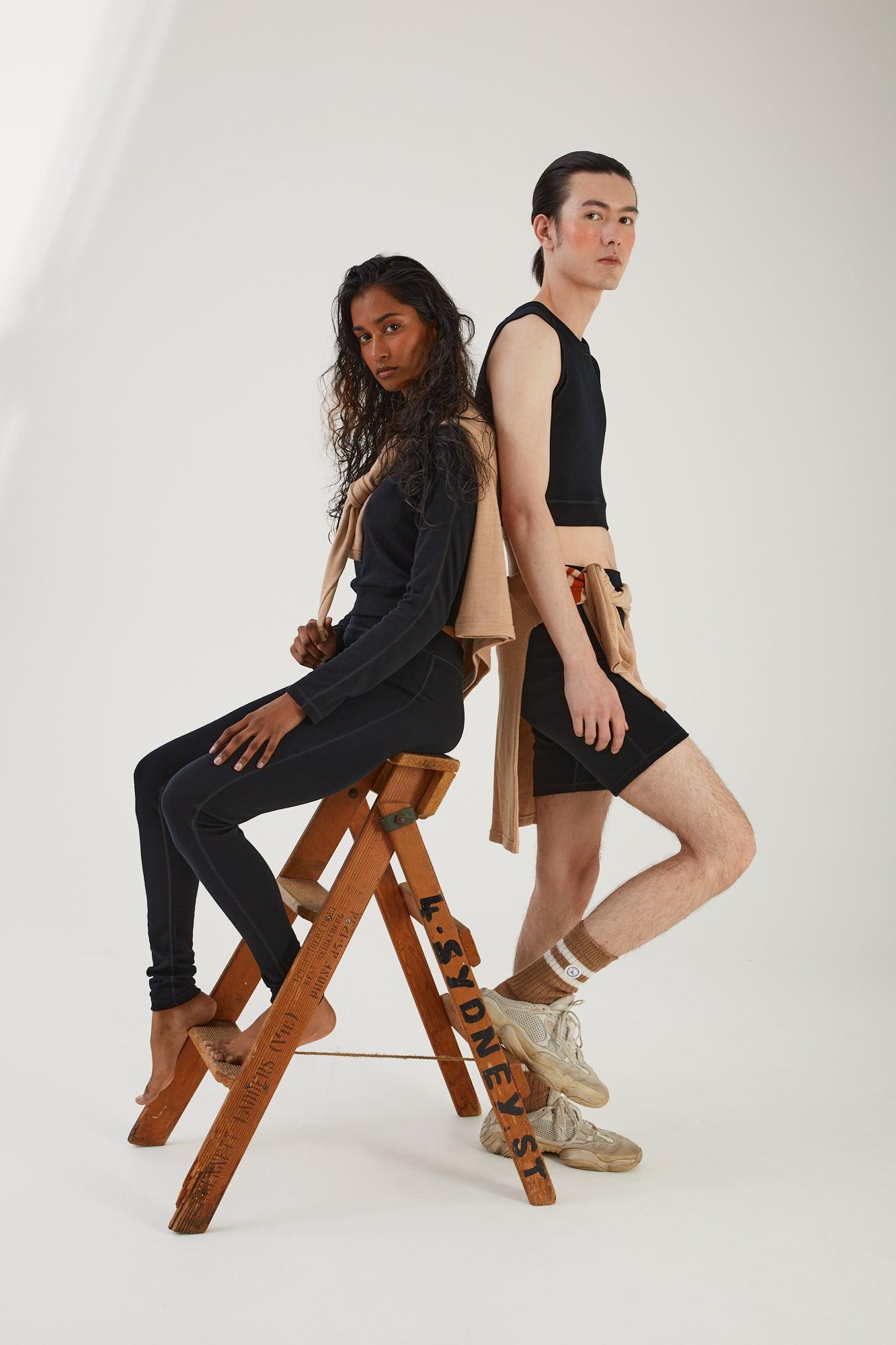 A person sitting on a wooden step ladder wearing merino wool leggings and a merino wool long sleeve cropped top with a merino wool sweater draped over their shoulders leaning against someone wearing merino wool socks, merino wool bike shorts and a merino wool cropped tank with a merino wool jumper wrapped around their waist