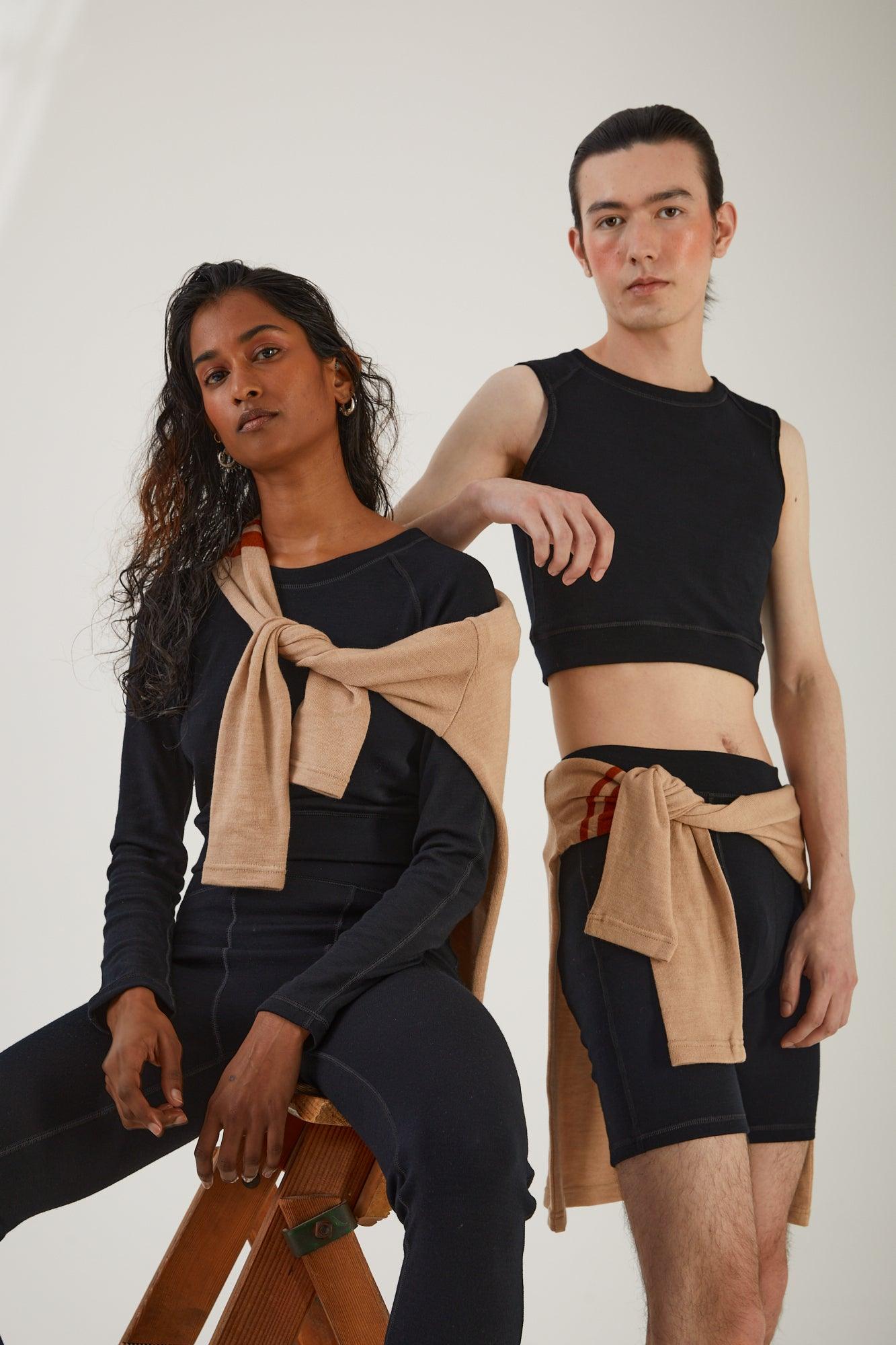 A person sitting on a wooden step ladder wearing merino wool leggings and a merino wool cropped long sleeve top with a merino wool jumper draped around their shoulders next to a person standing wearing merino wool biker shorts and merino wool cropped tank with a merino wool jumper tied around their waist